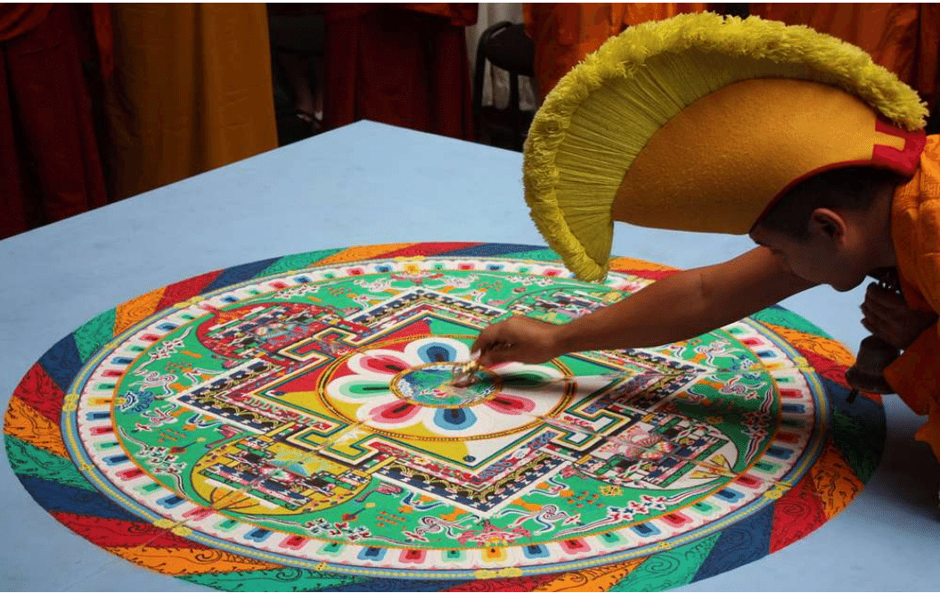 A Tibetan Buddhist monk in tall yellow hat applies finishing touches to a colourful sand mandala.